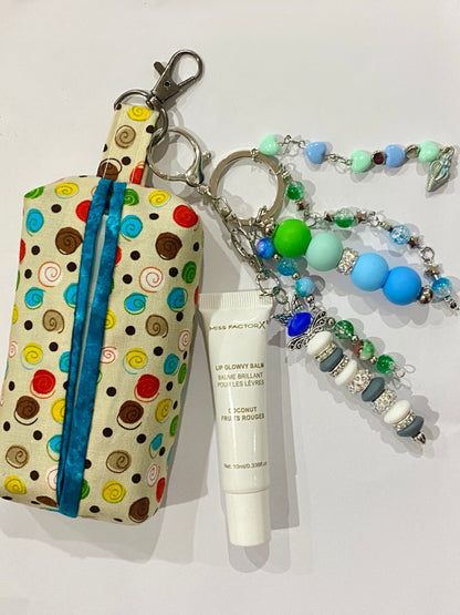 A fabric zippered pouch with a colorful polka dot pattern and a blue zipper, attached to a beaded keychain strap. A white tube of lip balm is placed in front of it on a plain white surface.